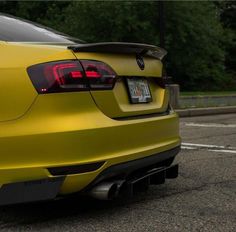 the rear end of a yellow car parked in a parking lot with trees in the background