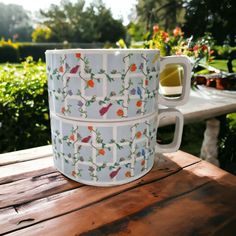 two mugs sitting on top of a wooden table in front of some bushes and flowers