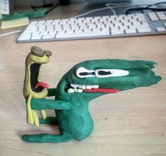 a clay model of a green dinosaur holding a man's head in front of a computer keyboard