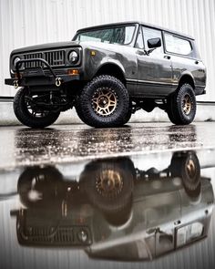 an suv parked in front of a building with its reflection on the wet ground next to it