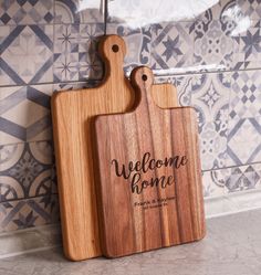 a wooden cutting board with the words welcome home on it next to a tiled wall