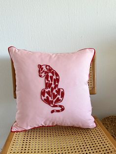 a pink pillow with a red cat embroidered on the front sitting on a wicker chair