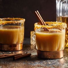 two glasses filled with drinks sitting on top of a table next to cinnamon sticks and an apple