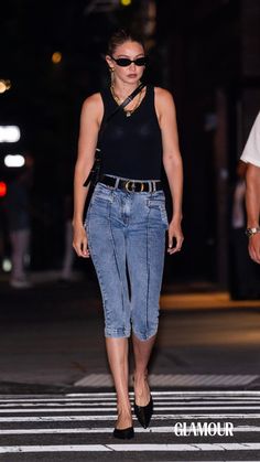 a woman in black top and jeans walking down the street at night with her hand on her hip