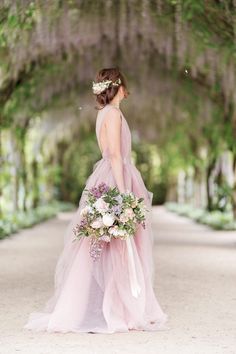 a woman in a pink dress is holding a bouquet and looking at the ground with trees behind her