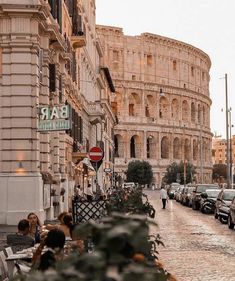 people are sitting at tables in front of the roman colossion, with cars parked on the street