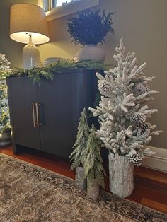 two christmas trees sitting on top of a wooden table next to a cabinet and lamp