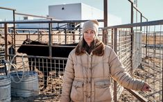 a woman standing in front of a fence with cows behind her and wearing a hat