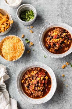 three bowls filled with chili and cheese on top of a gray table next to other dishes