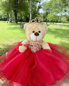 a teddy bear wearing a red dress and a tiara sitting on the ground in a park