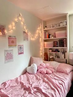 a bed with pink sheets and pillows in a room filled with bookshelves, lights and pictures on the wall