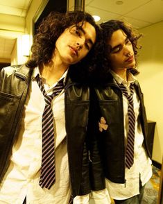 two young men wearing leather vests and ties in an office setting, one with long curly hair