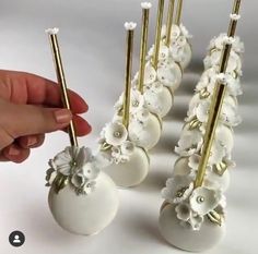 a hand is holding several white vases with flowers and gold sticks sticking out of them