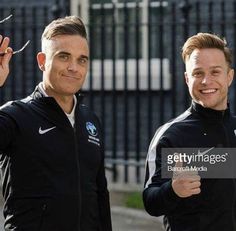 two men standing next to each other in front of a fence holding up their fingers