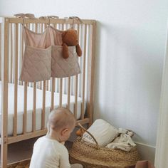 a baby sitting in front of a crib with a teddy bear hanging from it