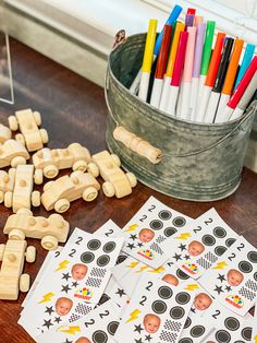 wooden toy cars and markers on a table next to a bucket of colored pencils