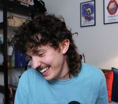 a man with curly hair laughing while holding a microphone in front of him and wearing a blue t - shirt