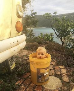 a baby sitting in a yellow bucket next to a car on the ground near water