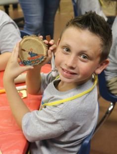 a young boy holding up a small wooden object at a table with other children around him