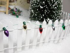 a white picket fence with christmas lights on it and a small tree in the background