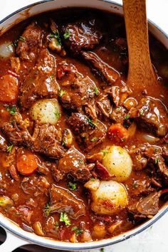 a pot full of beef stew with potatoes and carrots on the side next to a wooden spoon