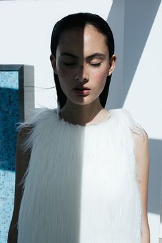 a woman with long black hair wearing a white feathered dress and standing in front of a wall