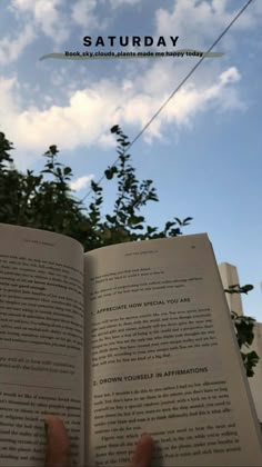 someone is holding an open book in front of a blue sky with clouds and trees