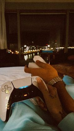 a person laying in bed holding a nintendo wii game controller with the city lights behind them