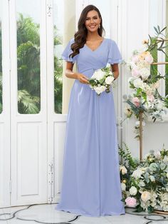 a woman in a long blue dress standing next to a flower arrangement and holding a bouquet