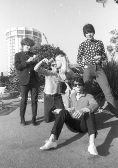 four people sitting on the ground in front of some bushes and trees with one person taking a selfie