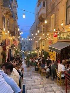 people sitting at tables in an alleyway with string lights strung above the walkways