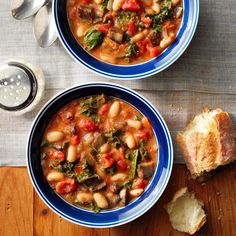 two bowls of soup on a wooden table with bread and spoons next to it