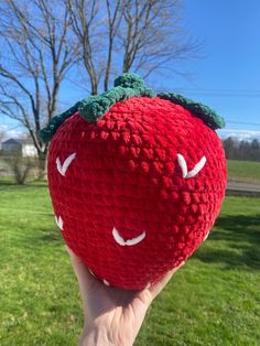 someone is holding up a crocheted strawberry in the grass with trees in the background