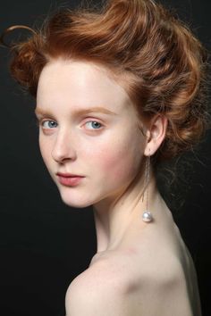 a woman with red hair and blue eyes is wearing pearls on her ear, looking at the camera