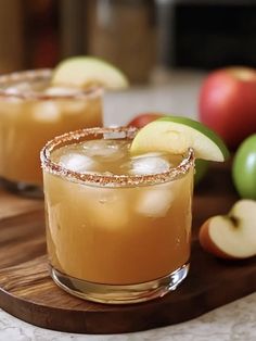 two glasses filled with apple cider sitting on top of a cutting board next to apples