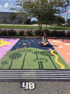 a woman sitting on the ground in front of a painted area with trees and flowers