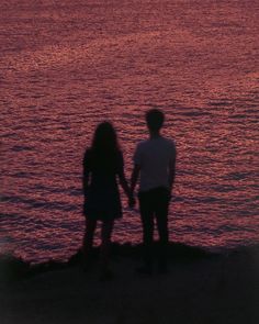 two people standing next to each other near the ocean at sunset or sunrise with an airplane flying in the sky