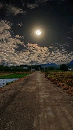 a dirt road under a cloudy sky with the moon in the distance and water below