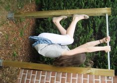 a woman laying on top of a wooden bench next to a green bush and shrubbery