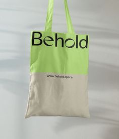 a green and white tote bag hanging from a hook with the word behold printed on it