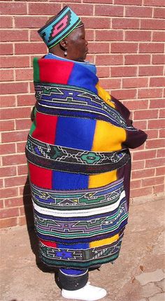 a man wearing a multicolored jacket and hat standing in front of a brick wall