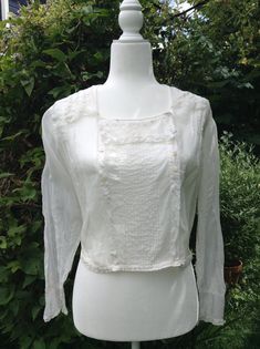 Antique from the 1910s, this is not a reproduction. Features pin tucks and decorative buttons down the front bib with a buttoned side closure. Square neckline with delicate embroidery and lace trim on the collar. Lightweight white cotton. Long sleeves with lace trim around single button cuffs. One of the decorative buttons is broken on the front and there is a small hole near the right cuff. Good condition for its age. Material: Cotton Size: S US Measurements *All measurements are approximate an Victorian Fitted Tops For Summer, Fitted Victorian Summer Tops, Fitted Victorian Tops For Summer, Victorian Style Daywear Blouse With Buttons, Victorian Blouse With Buttons For Daywear, Victorian Style Blouse With Buttons For Daywear, Fitted Vintage Blouse For Gatherings, Victorian Style Blouse For Daywear With Buttons, Victorian Long Sleeve Tops With Buttons