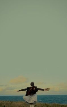 a woman standing on top of a grass covered field next to the ocean with her arms outstretched