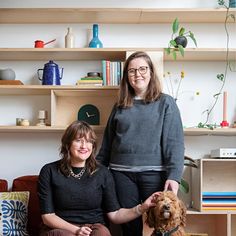 two women sitting on a couch with a dog in front of them and bookshelves behind them