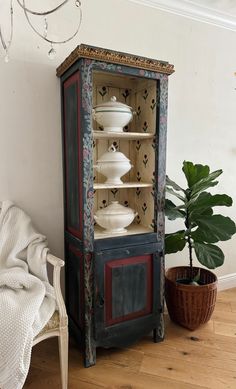 an old china cabinet with dishes on it in a living room next to a potted plant