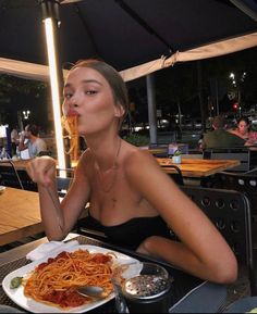 a woman sitting at an outdoor table eating spaghetti