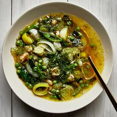 a white bowl filled with soup and vegetables on top of a wooden table next to a spoon