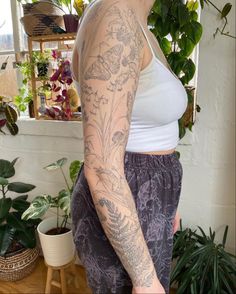 a woman with tattoos on her arm and leg, standing in front of potted plants