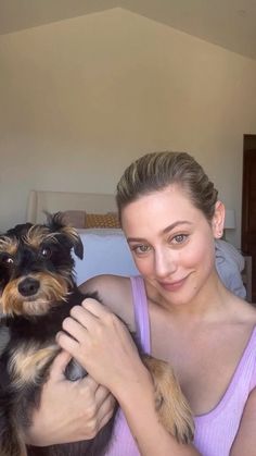 a woman is holding a small dog in her arms and smiling at the camera while sitting on a bed