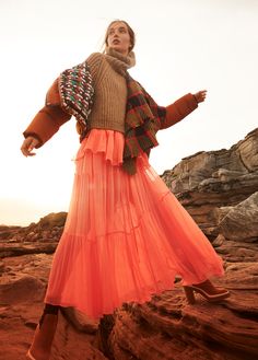 a woman in an orange skirt and sweater standing on rocks